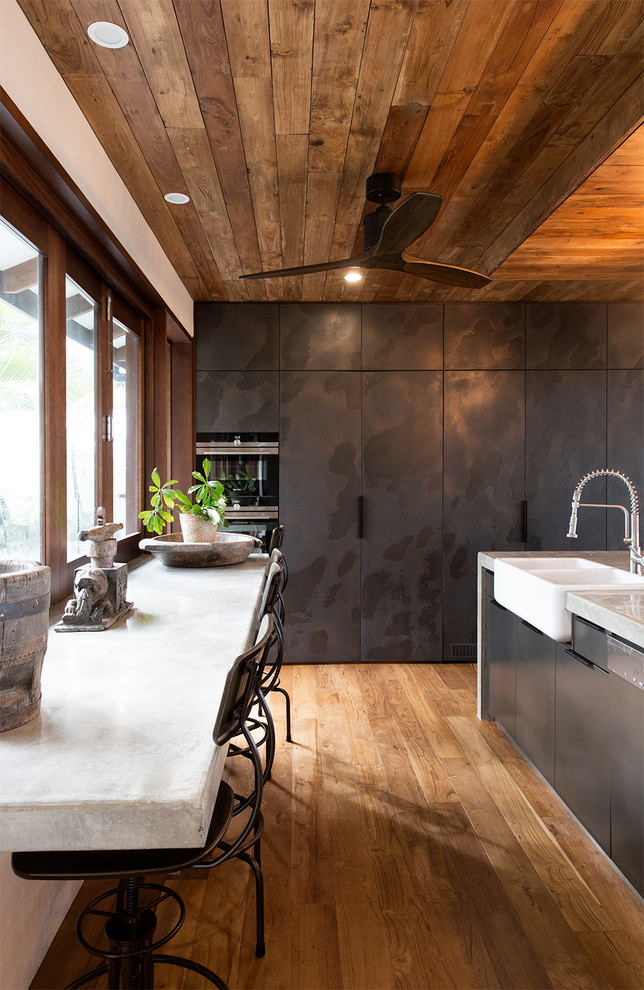 Cette photo montre une cuisine bicolore exotique avec un évier de ferme, un placard à porte plane, des portes de placard noires, un sol en bois brun, îlot et un sol marron.