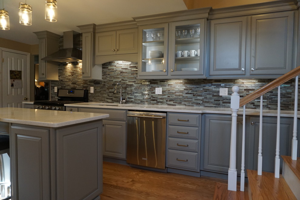 This is an example of a small traditional l-shaped kitchen/diner in Providence with a triple-bowl sink, raised-panel cabinets, grey cabinets, engineered stone countertops, grey splashback, mosaic tiled splashback, stainless steel appliances, medium hardwood flooring, an island and white worktops.