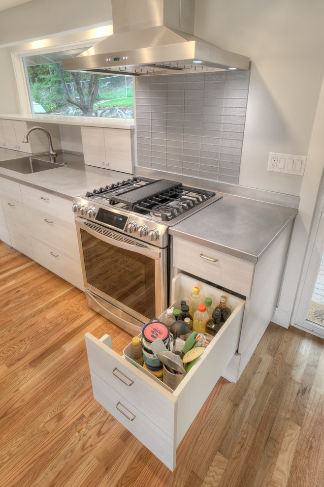 This is an example of a small contemporary l-shaped open plan kitchen in Seattle with a single-bowl sink, flat-panel cabinets, stainless steel worktops, grey splashback, ceramic splashback, stainless steel appliances, light hardwood flooring, an island and white cabinets.