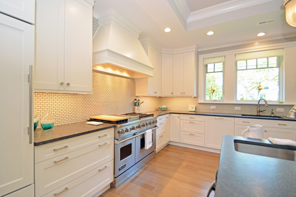 Bild på ett vintage kök, med en undermonterad diskho, luckor med infälld panel, vita skåp, granitbänkskiva, vitt stänkskydd, stänkskydd i tunnelbanekakel, integrerade vitvaror, ljust trägolv och en köksö