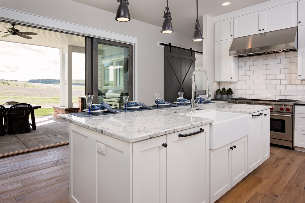 Modelo de cocina de estilo de casa de campo, grande, abierta, con fregadero sobremueble, armarios estilo shaker, puertas de armario blancas, encimera de granito, salpicadero blanco, salpicadero de azulejos tipo metro, electrodomésticos de acero inoxidable, suelo de madera clara y una isla