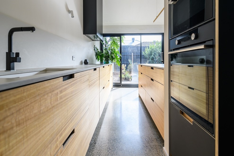 Large modern galley kitchen/diner in Melbourne with a submerged sink, flat-panel cabinets, light wood cabinets, engineered stone countertops, grey splashback, stone slab splashback, concrete flooring, an island, white worktops, black appliances and grey floors.
