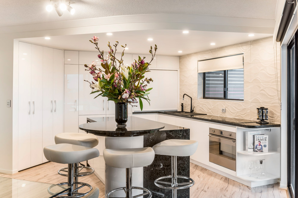 Photo of a medium sized contemporary l-shaped kitchen/diner in Brisbane with a built-in sink, flat-panel cabinets, white cabinets, granite worktops, beige splashback, cement tile splashback, stainless steel appliances, light hardwood flooring and an island.