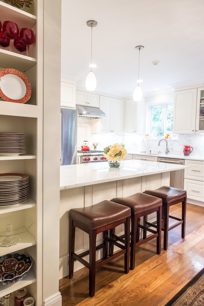 Klassisk inredning av ett mellanstort kök, med luckor med upphöjd panel, vita skåp, vitt stänkskydd, rostfria vitvaror, en halv köksö, en undermonterad diskho, ljust trägolv, marmorbänkskiva och stänkskydd i stenkakel