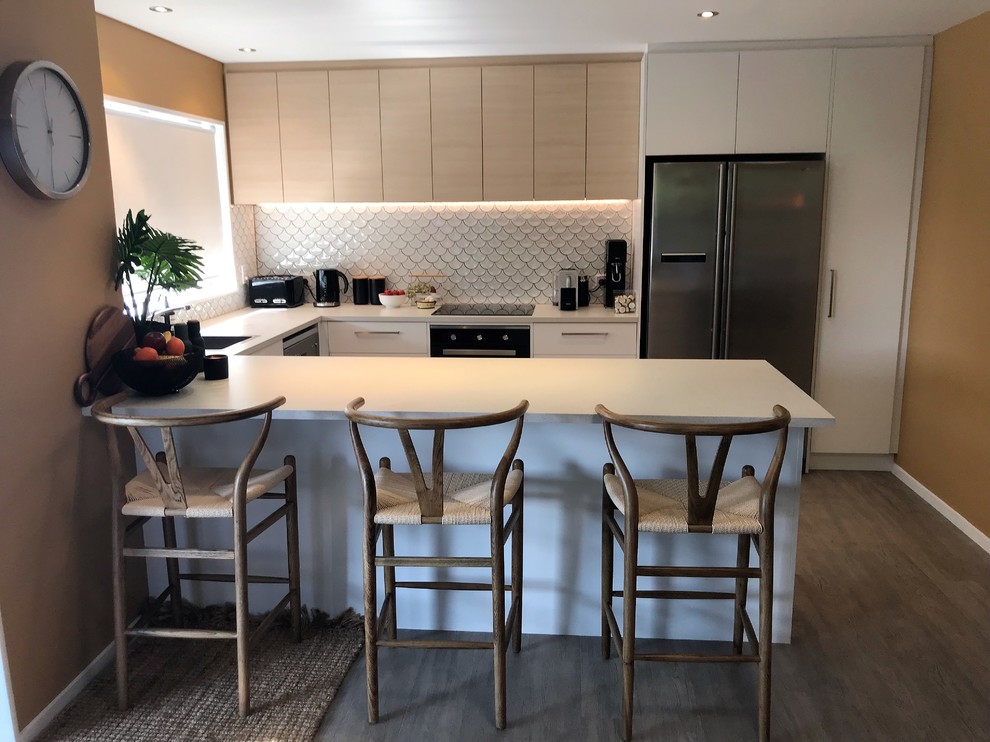 Example of a mid-sized trendy u-shaped vinyl floor and gray floor open concept kitchen design in Christchurch with a single-bowl sink, flat-panel cabinets, white cabinets, laminate countertops, white backsplash, ceramic backsplash, stainless steel appliances and white countertops