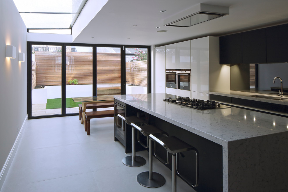 Design ideas for a large contemporary single-wall open plan kitchen in London with an integrated sink, flat-panel cabinets, white cabinets, granite worktops, integrated appliances, ceramic flooring, a breakfast bar, white floors, black splashback and glass sheet splashback.