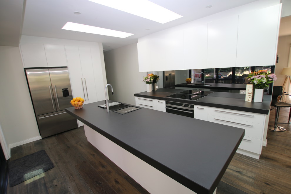 This is an example of a large l-shaped kitchen pantry in Sydney with a submerged sink, flat-panel cabinets, white cabinets, tile countertops, black splashback, mirror splashback, stainless steel appliances, medium hardwood flooring and an island.