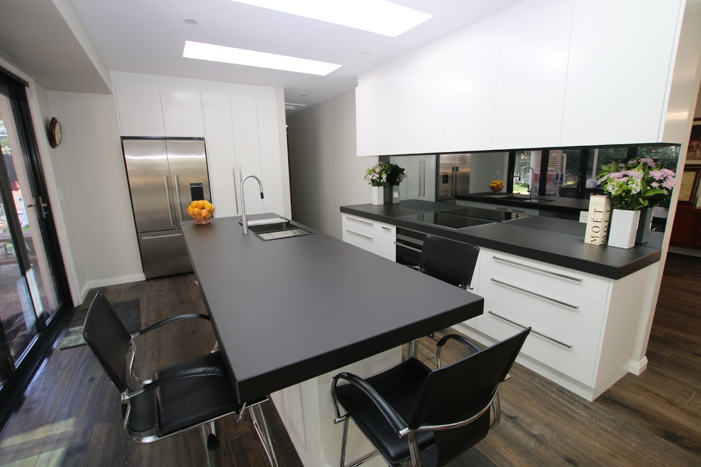 Exempel på ett stort kök, med en undermonterad diskho, släta luckor, vita skåp, kaklad bänkskiva, svart stänkskydd, spegel som stänkskydd, rostfria vitvaror, mellanmörkt trägolv och en köksö