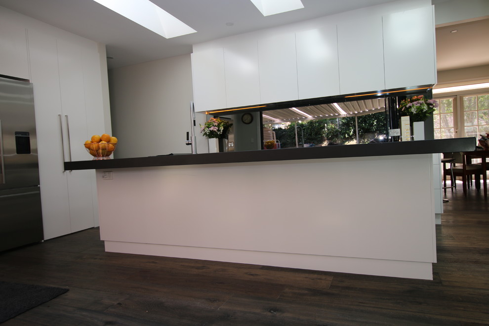 Bild på ett stort kök, med en undermonterad diskho, släta luckor, vita skåp, kaklad bänkskiva, svart stänkskydd, spegel som stänkskydd, rostfria vitvaror, mellanmörkt trägolv och en köksö