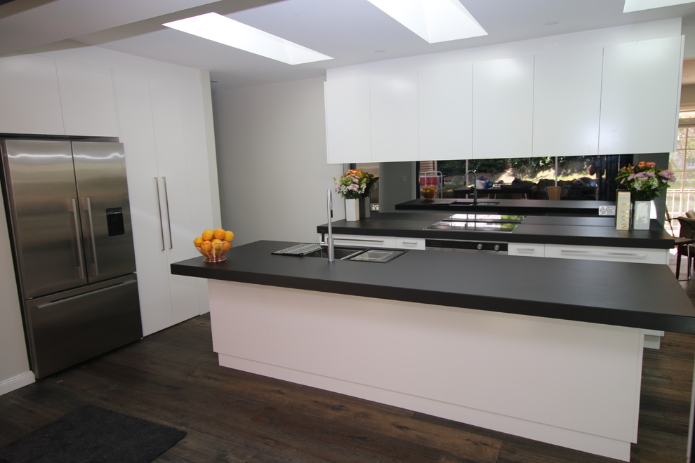 Example of a large l-shaped medium tone wood floor kitchen pantry design in Sydney with an undermount sink, flat-panel cabinets, white cabinets, tile countertops, black backsplash, mirror backsplash, stainless steel appliances and an island