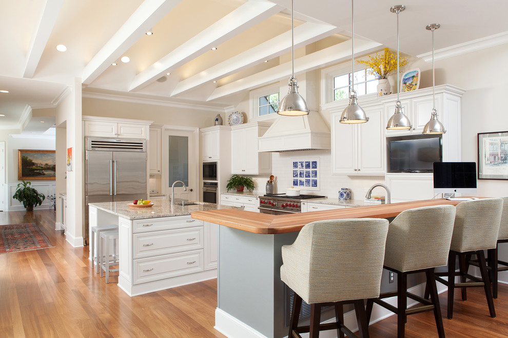Landhausstil Küche in L-Form mit profilierten Schrankfronten, weißen Schränken, Küchenrückwand in Weiß, Küchengeräten aus Edelstahl, braunem Holzboden und Kücheninsel in Sonstige