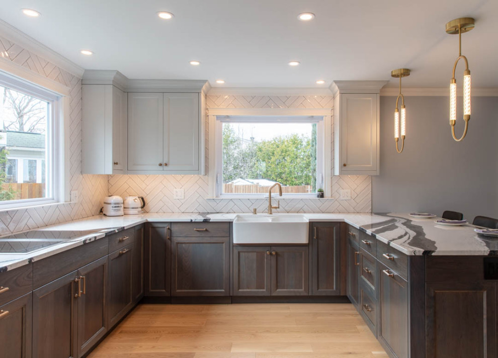Photo of a traditional u-shaped open plan kitchen in Ottawa with a belfast sink, recessed-panel cabinets, dark wood cabinets, engineered stone countertops, white splashback, porcelain splashback, integrated appliances, light hardwood flooring, a breakfast bar and black worktops.