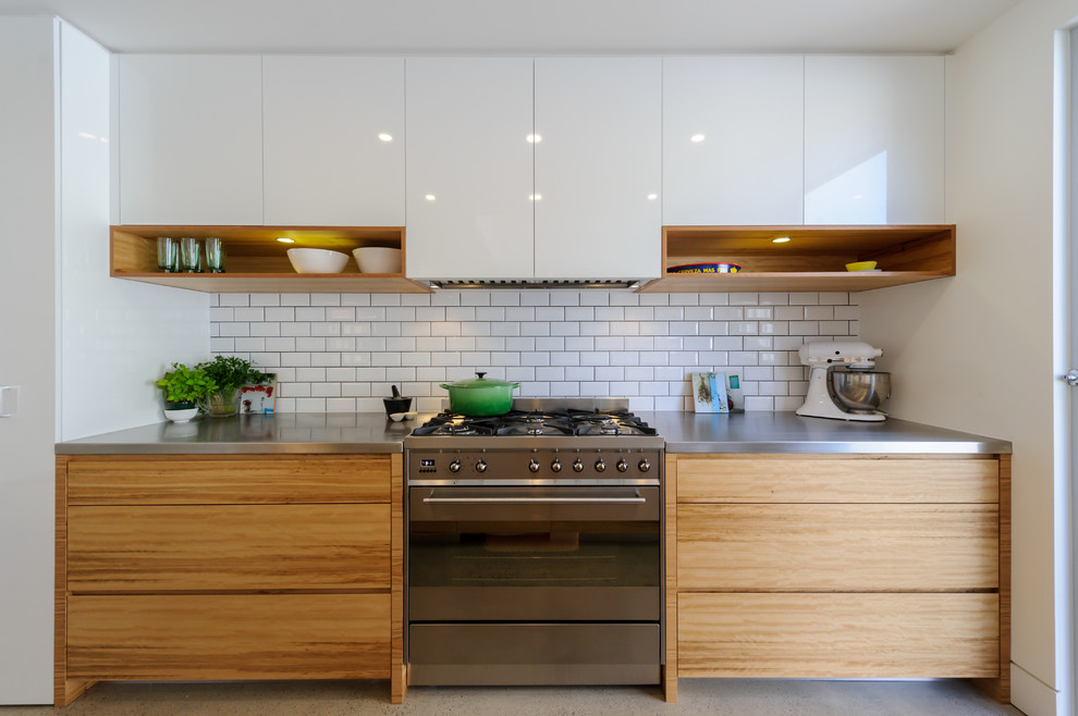 Moderne Küche mit flächenbündigen Schrankfronten, hellbraunen Holzschränken, Edelstahl-Arbeitsplatte, Küchenrückwand in Weiß, Rückwand aus Metrofliesen und Küchengeräten aus Edelstahl in Melbourne