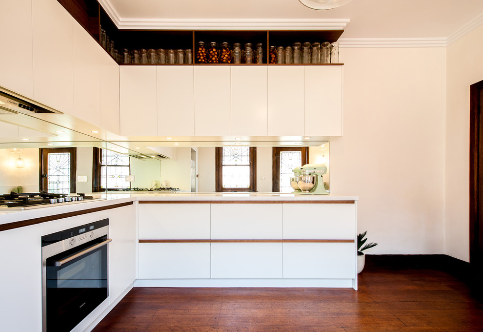 Geschlossene Moderne Küche ohne Insel in L-Form mit Unterbauwaschbecken, flächenbündigen Schrankfronten, weißen Schränken, Quarzwerkstein-Arbeitsplatte, Glasrückwand, Küchengeräten aus Edelstahl und braunem Holzboden in Melbourne