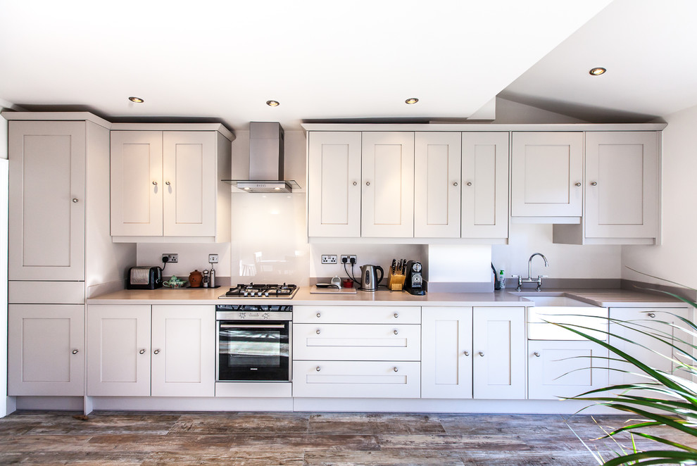 Medium sized modern single-wall kitchen/diner in London with a belfast sink, shaker cabinets, beige cabinets, composite countertops, grey splashback, stone slab splashback, integrated appliances, painted wood flooring and no island.