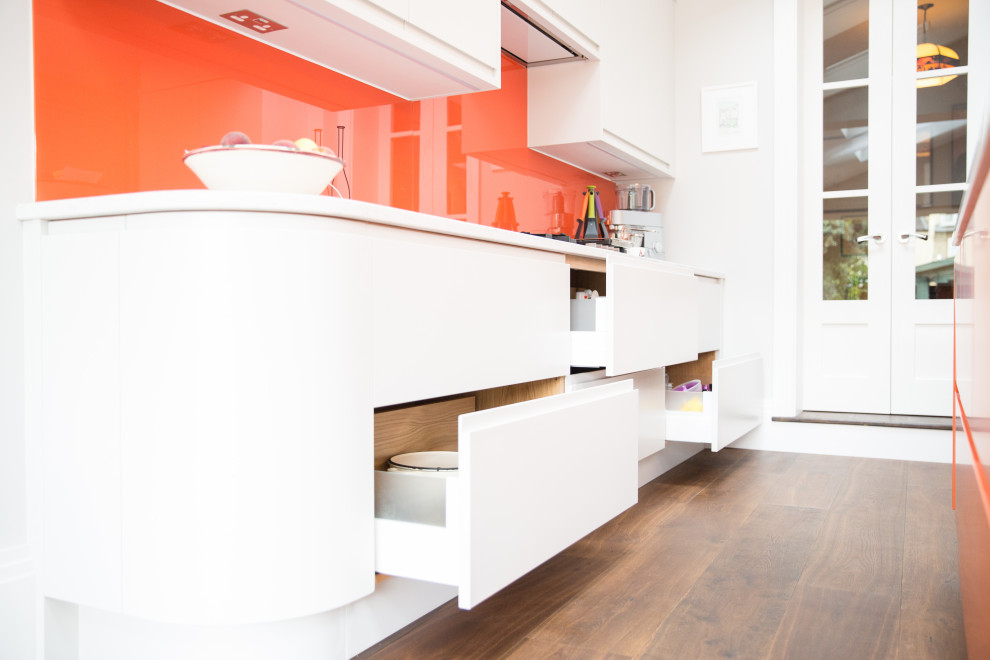 Inredning av ett modernt mellanstort vit vitt kök, med en enkel diskho, släta luckor, orange skåp, bänkskiva i koppar, orange stänkskydd, stänkskydd i glaskakel, mörkt trägolv, en köksö och brunt golv