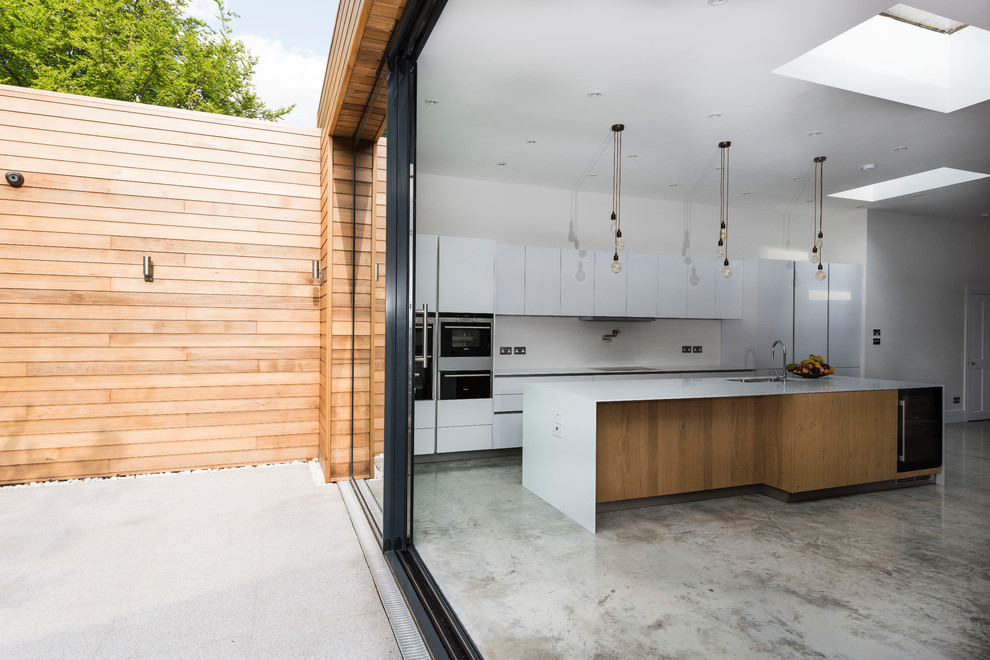 This is an example of a large contemporary kitchen/diner in London with a double-bowl sink, white cabinets, composite countertops, stainless steel appliances, concrete flooring and an island.