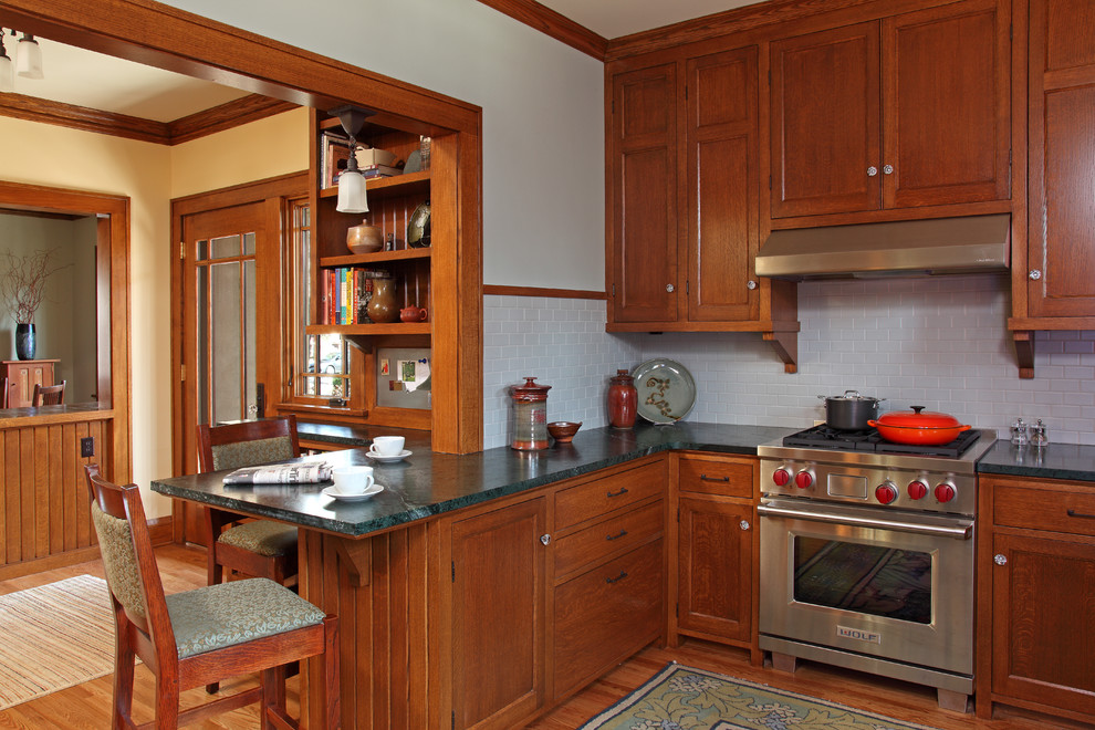 Bild på ett amerikanskt kök, med skåp i mellenmörkt trä, luckor med infälld panel, stänkskydd i tunnelbanekakel, rostfria vitvaror, vitt stänkskydd, en rustik diskho, bänkskiva i täljsten, en halv köksö och mellanmörkt trägolv