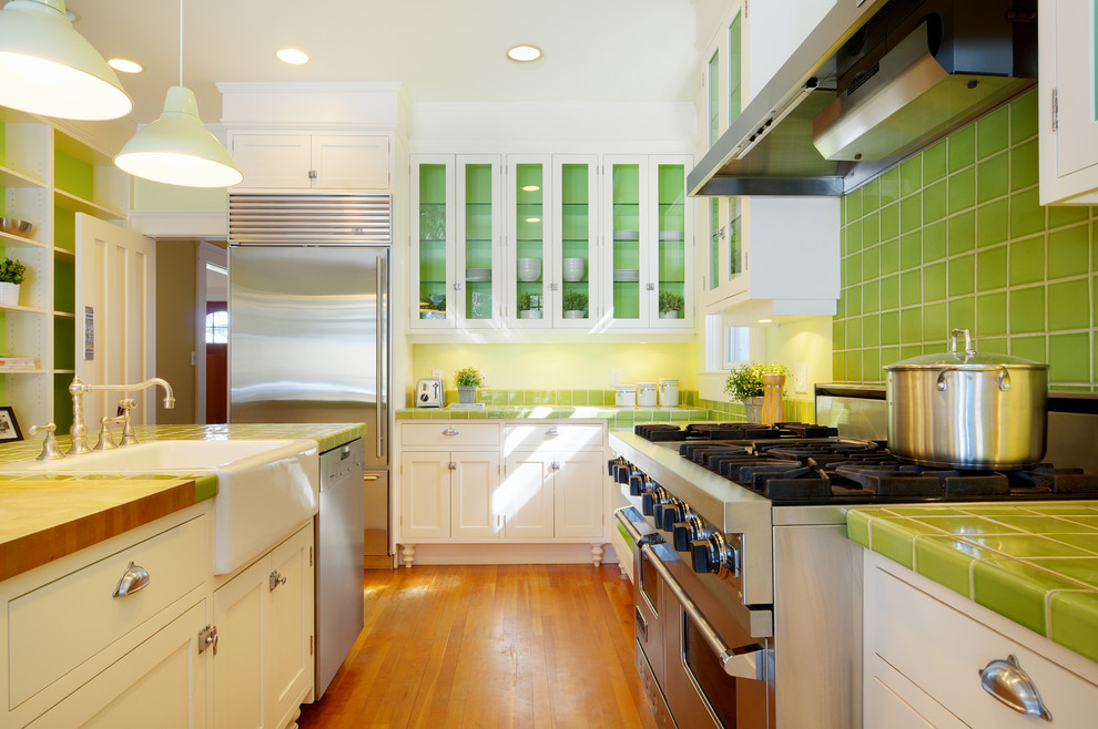 Exemple d'une cuisine chic avec un électroménager en acier inoxydable, un évier de ferme, plan de travail carrelé, un placard à porte vitrée, des portes de placard blanches, une crédence verte et un plan de travail vert.