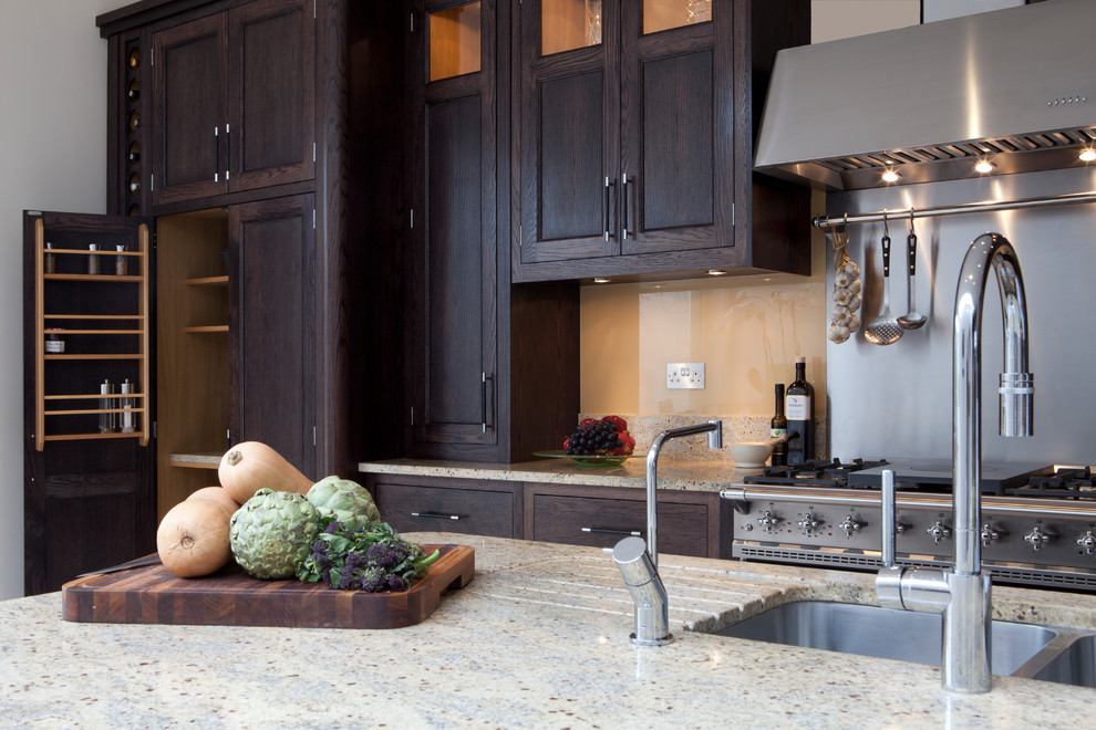 Bild på ett stort vintage linjärt kök och matrum, med en integrerad diskho, luckor med infälld panel, svarta skåp, granitbänkskiva, beige stänkskydd, glaspanel som stänkskydd, rostfria vitvaror, kalkstensgolv och en köksö