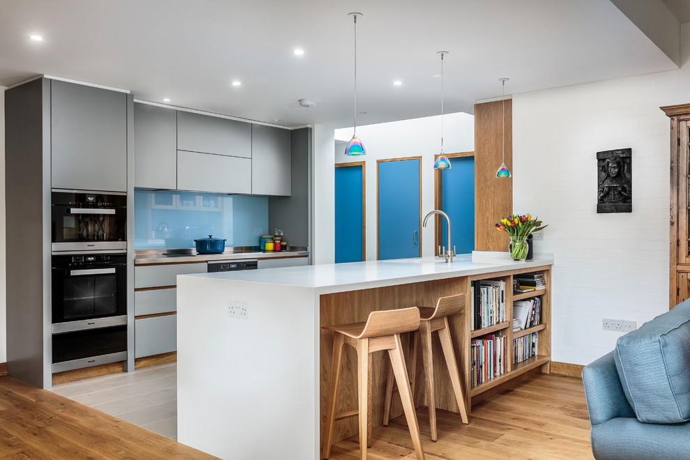 Идея дизайна: параллельная кухня в современном стиле с плоскими фасадами, серыми фасадами, синим фартуком, фартуком из стекла, черной техникой, полуостровом, бежевым полом и белой столешницей
