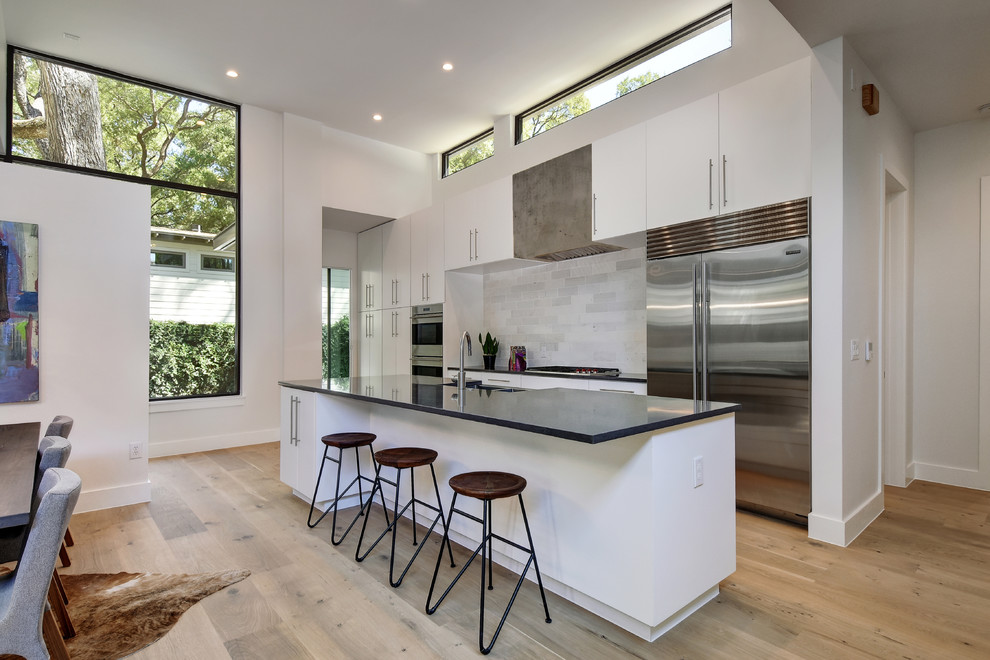 Design ideas for a contemporary galley kitchen/diner in Austin with flat-panel cabinets, white cabinets, grey splashback, stainless steel appliances, light hardwood flooring and an island.