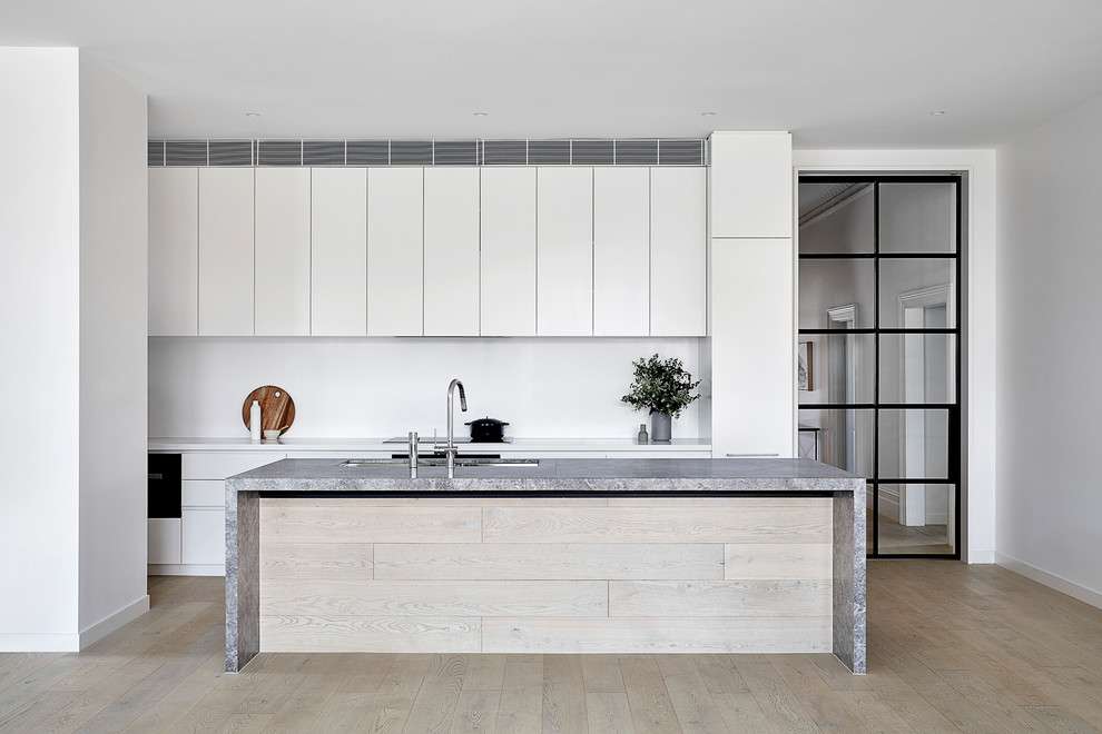 Contemporary galley kitchen in Melbourne with a submerged sink, flat-panel cabinets, white cabinets, white splashback, light hardwood flooring, an island, beige floors and grey worktops.