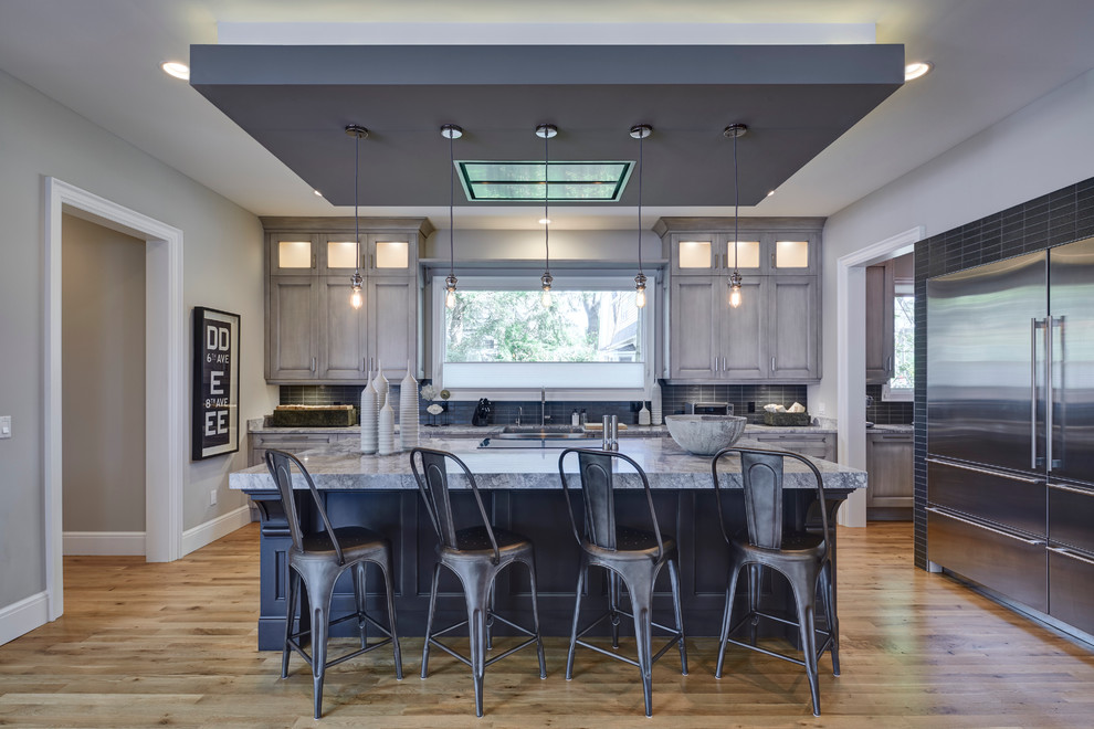 This is an example of a large urban l-shaped open plan kitchen in Detroit with quartz worktops, grey splashback, glass tiled splashback, stainless steel appliances, an island, light hardwood flooring, distressed cabinets, a belfast sink and recessed-panel cabinets.