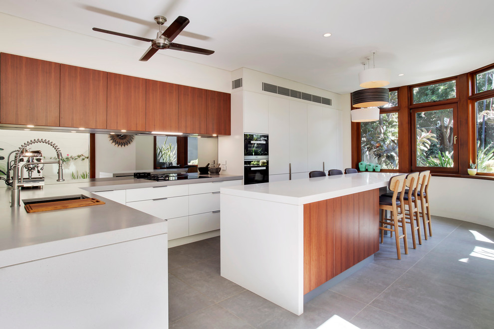 Modern inredning av ett stort kök, med en undermonterad diskho, släta luckor, vita skåp, bänkskiva i kvarts, grått stänkskydd, spegel som stänkskydd, svarta vitvaror, betonggolv och en köksö