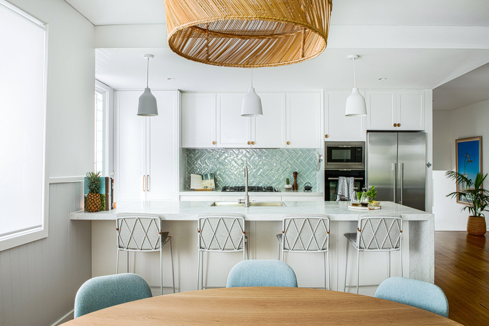 Idées déco pour une cuisine américaine bord de mer avec un placard à porte shaker, des portes de placard blanches, une crédence verte, un électroménager en acier inoxydable, un sol en bois brun, îlot et un plan de travail blanc.