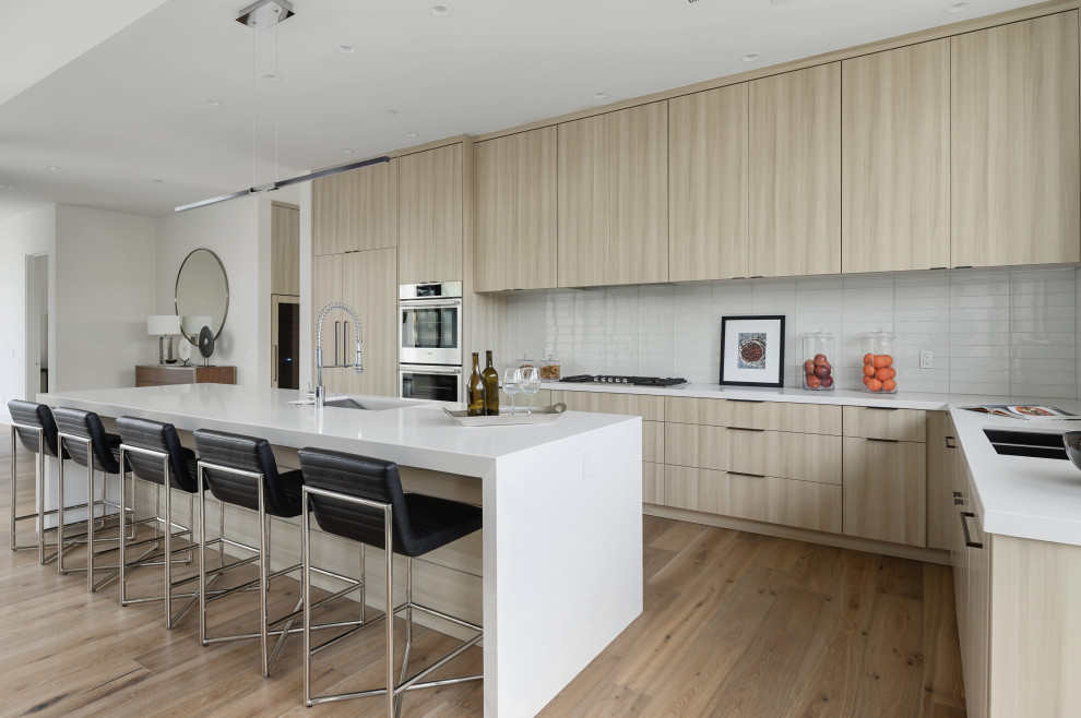 Example of a trendy l-shaped medium tone wood floor and brown floor kitchen design in Other with an undermount sink, flat-panel cabinets, light wood cabinets, white backsplash, stainless steel appliances, an island and white countertops