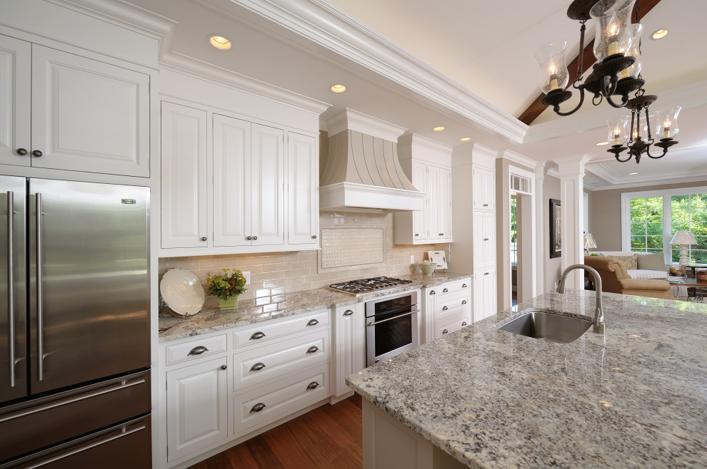 Example of a classic medium tone wood floor and tray ceiling kitchen design with an undermount sink, flat-panel cabinets, white cabinets, beige backsplash, subway tile backsplash, stainless steel appliances and an island