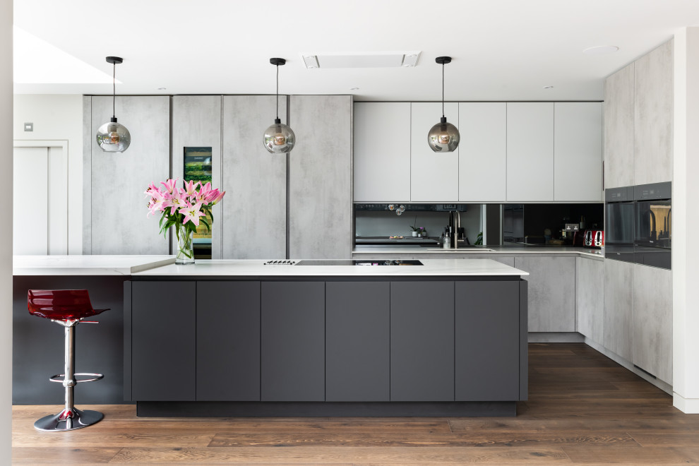Inspiration for an expansive contemporary l-shaped open plan kitchen in Surrey with a built-in sink, grey cabinets, quartz worktops, mirror splashback, stainless steel appliances, medium hardwood flooring, an island, brown floors and white worktops.