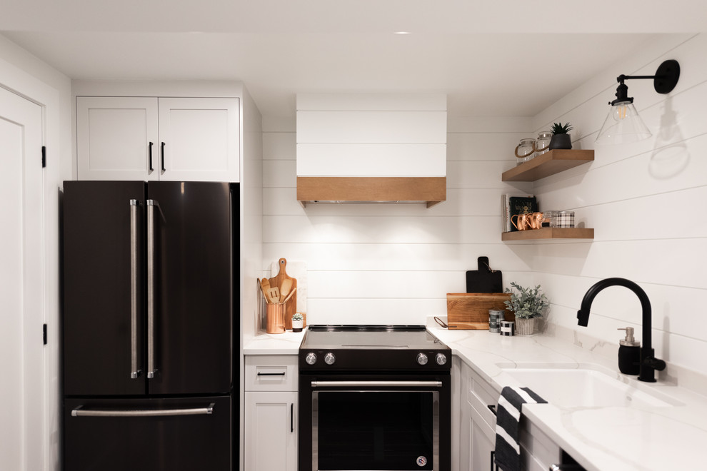 This is an example of a small rural l-shaped open plan kitchen in Montreal with a single-bowl sink, shaker cabinets, white cabinets, quartz worktops, white splashback, wood splashback, black appliances, no island, grey floors, white worktops and laminate floors.