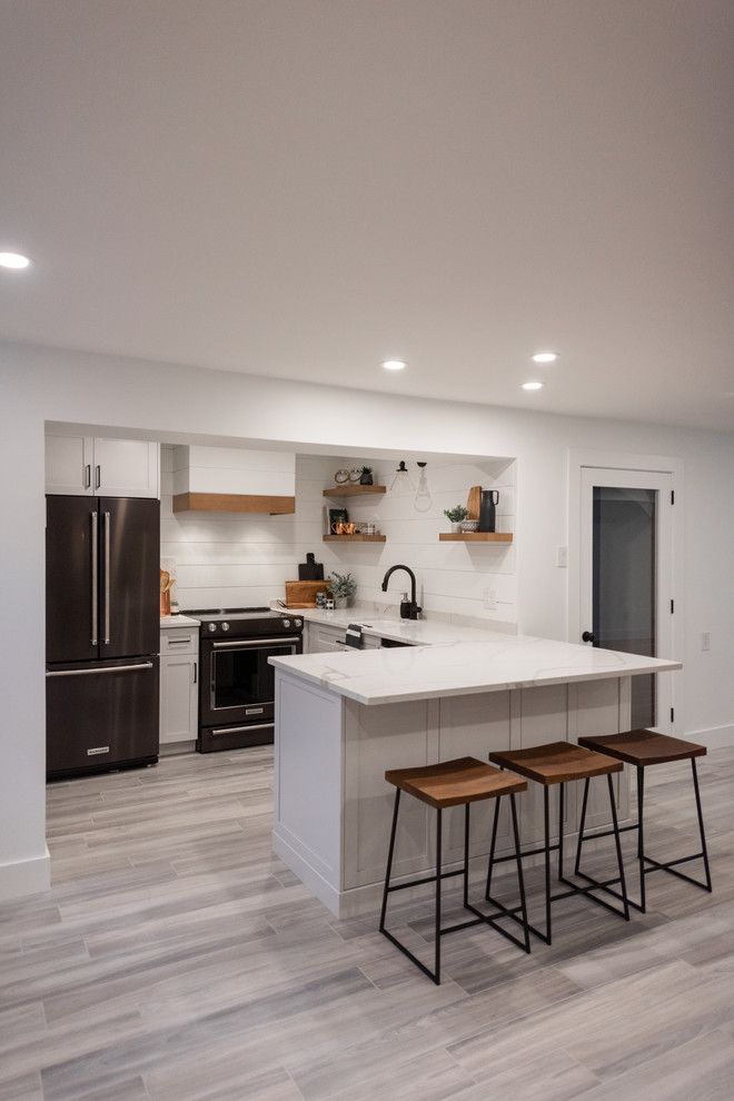 Example of a small farmhouse l-shaped laminate floor and gray floor open concept kitchen design in Montreal with a single-bowl sink, shaker cabinets, white cabinets, quartzite countertops, white backsplash, wood backsplash, black appliances, no island and white countertops