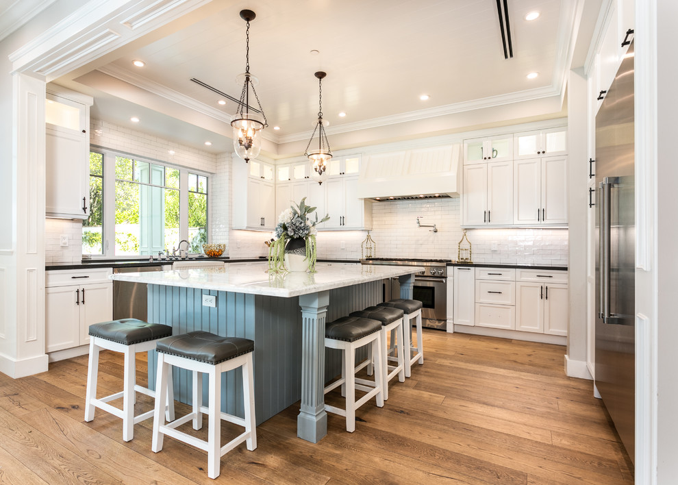 Photo of a traditional u-shaped kitchen in Los Angeles with a belfast sink, shaker cabinets, white cabinets, white splashback, metro tiled splashback, stainless steel appliances, medium hardwood flooring, an island, brown floors and black worktops.