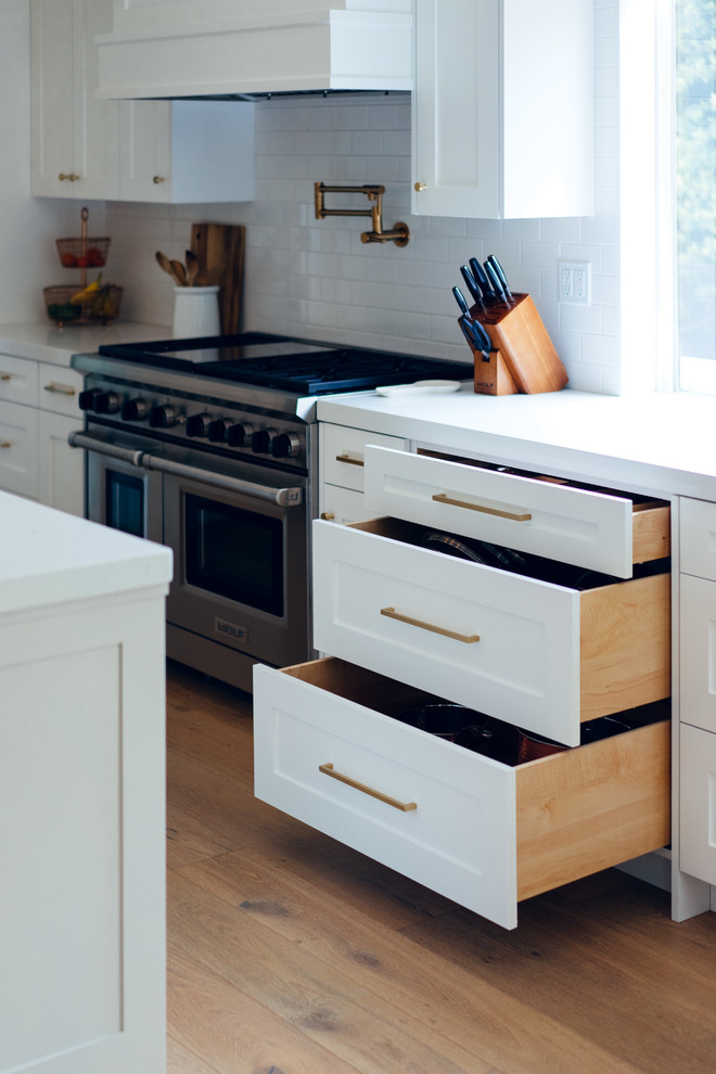 Photo of a large contemporary l-shaped kitchen/diner in Sacramento with a belfast sink, shaker cabinets, white cabinets, engineered stone countertops, white splashback, metro tiled splashback, stainless steel appliances, medium hardwood flooring, an island, brown floors and white worktops.