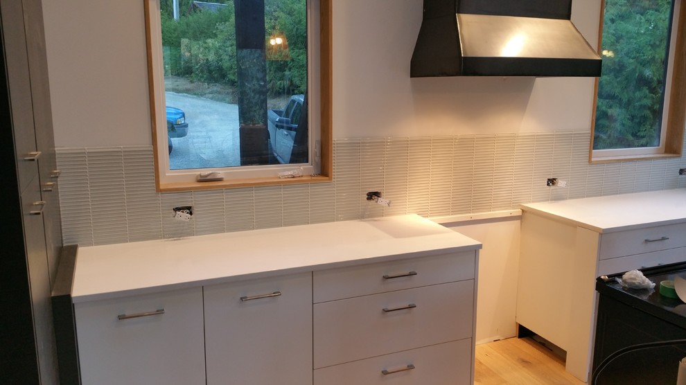 This is an example of a medium sized modern l-shaped open plan kitchen in Vancouver with a submerged sink, flat-panel cabinets, white cabinets, granite worktops, white splashback, glass tiled splashback, stainless steel appliances, light hardwood flooring and an island.