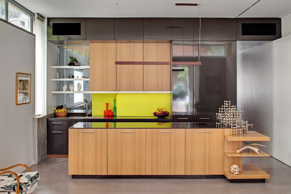 На фото: кухня-гостиная в современном стиле с одинарной мойкой, плоскими фасадами, светлыми деревянными фасадами, зеленым фартуком, бетонным полом, островом, серым полом, коричневой столешницей и двухцветным гарнитуром