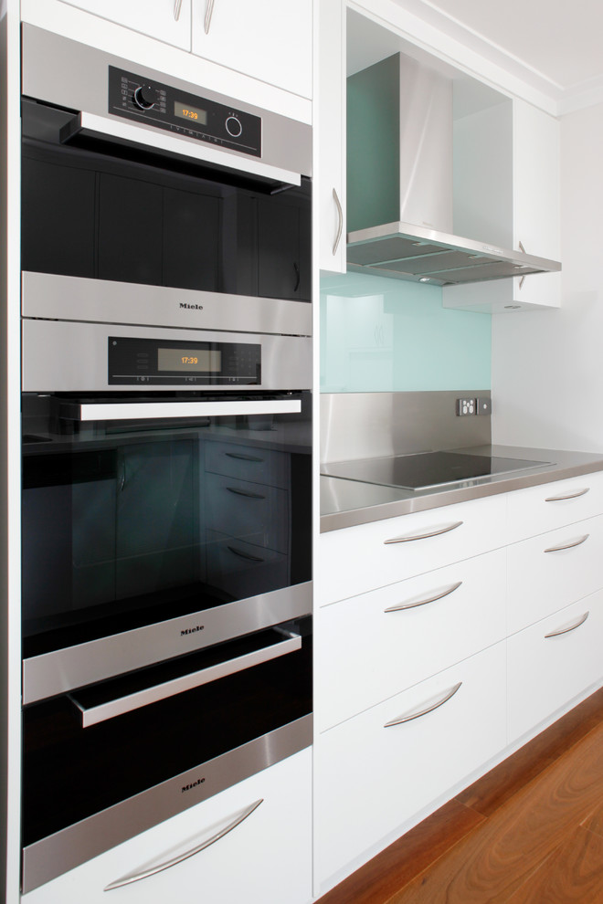 Modelo de cocina moderna con armarios con paneles lisos, puertas de armario blancas, encimera de acero inoxidable, salpicadero azul, salpicadero de vidrio templado y electrodomésticos de acero inoxidable