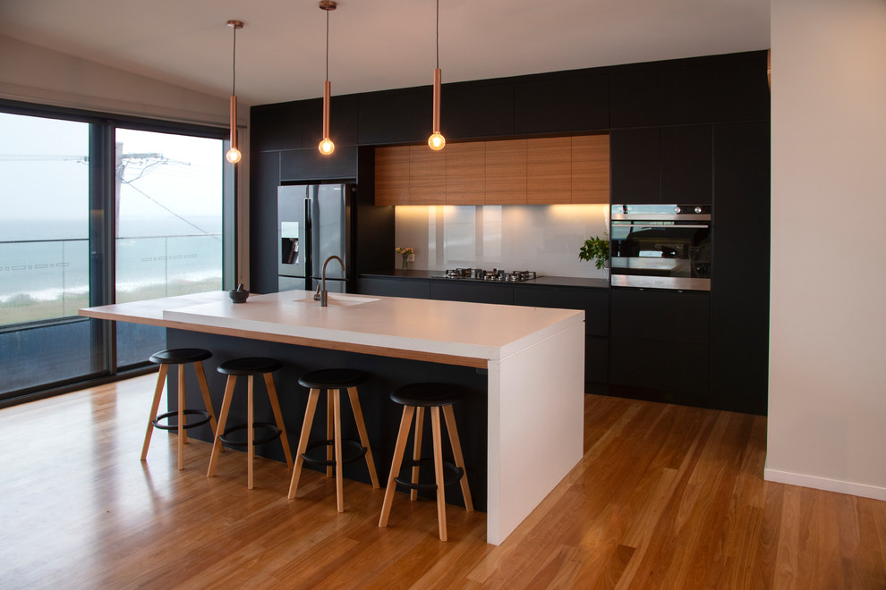 Inspiration for a medium sized contemporary galley open plan kitchen in Newcastle - Maitland with a submerged sink, flat-panel cabinets, black cabinets, concrete worktops, white splashback, glass sheet splashback, stainless steel appliances, medium hardwood flooring, an island, brown floors and grey worktops.