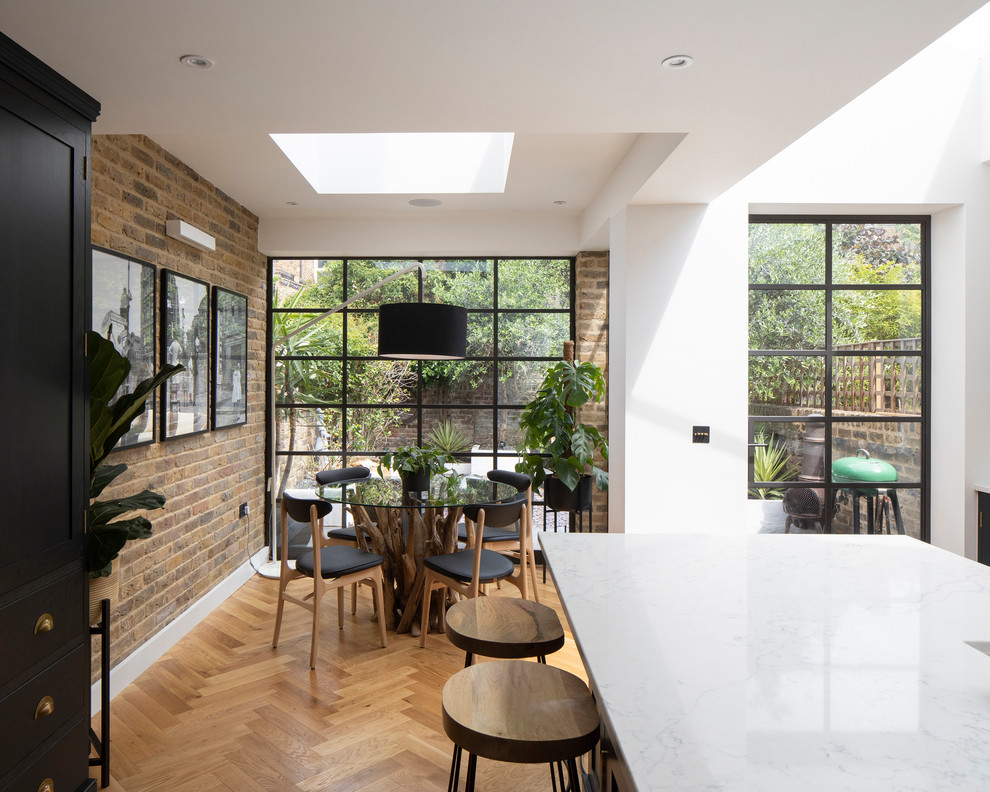 Inspiration for an eclectic galley kitchen/diner in London with a built-in sink, recessed-panel cabinets, black cabinets, marble worktops, brick splashback, black appliances, light hardwood flooring, an island, brown floors and white worktops.