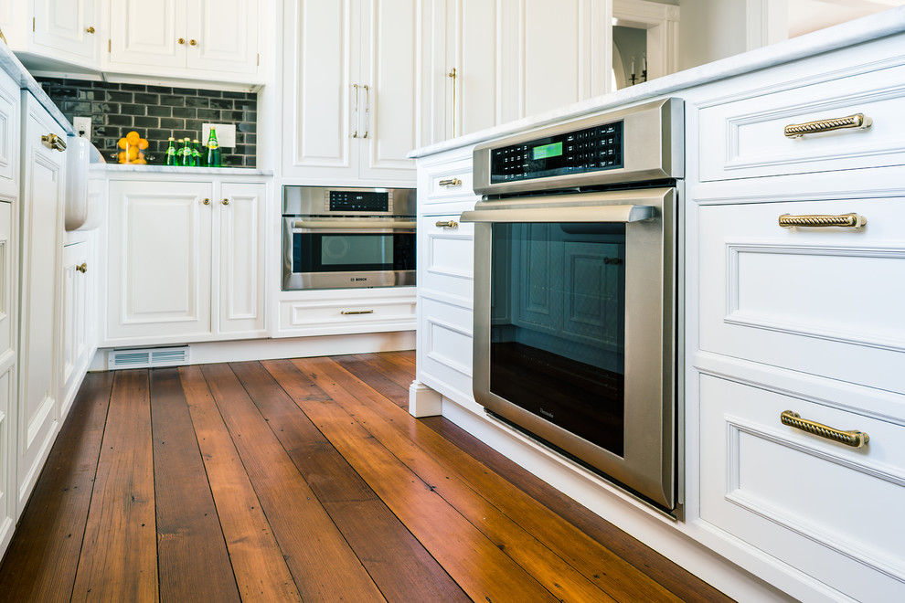 Example of a mid-sized transitional l-shaped medium tone wood floor eat-in kitchen design in Philadelphia with a farmhouse sink, glass-front cabinets, white cabinets, marble countertops, gray backsplash, ceramic backsplash, stainless steel appliances and an island