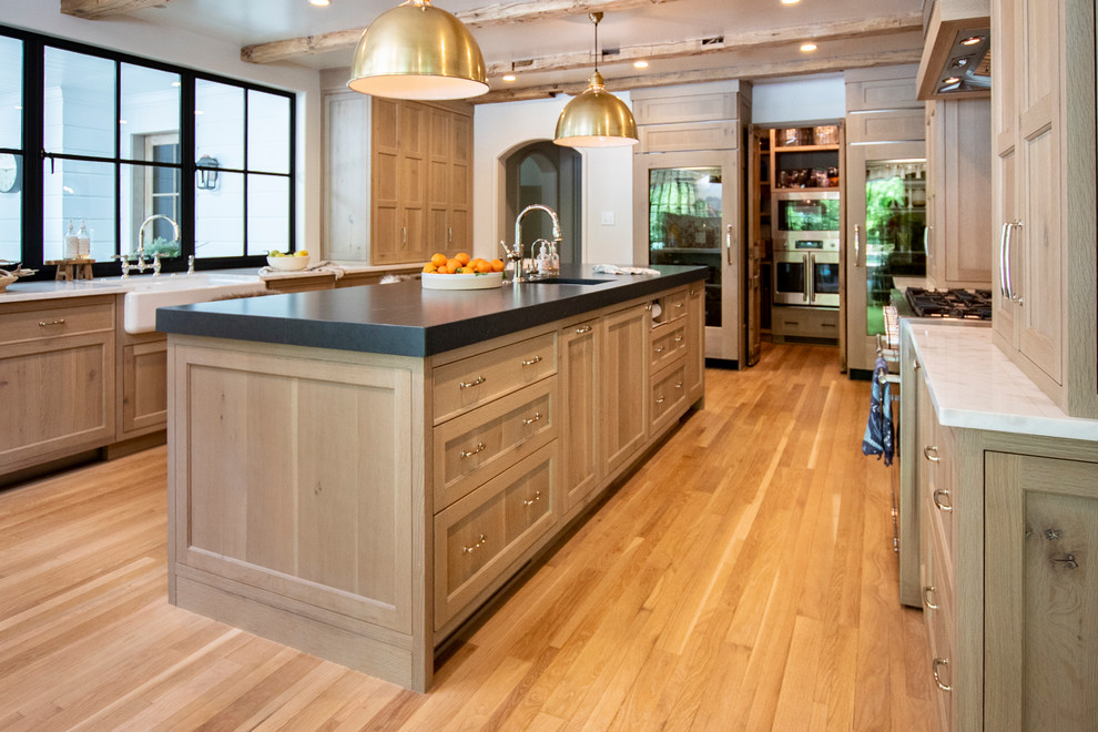 Mid-sized country u-shaped light wood floor and brown floor eat-in kitchen photo in Other with a farmhouse sink, shaker cabinets, light wood cabinets, quartz countertops, white backsplash, stone slab backsplash, paneled appliances, an island and black countertops
