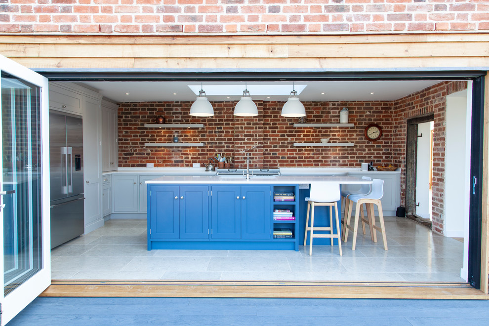 Cette image montre une cuisine traditionnelle en U fermée avec un placard à porte shaker, des portes de placard bleues, une crédence en brique, un électroménager en acier inoxydable, îlot, un sol blanc et un plan de travail blanc.
