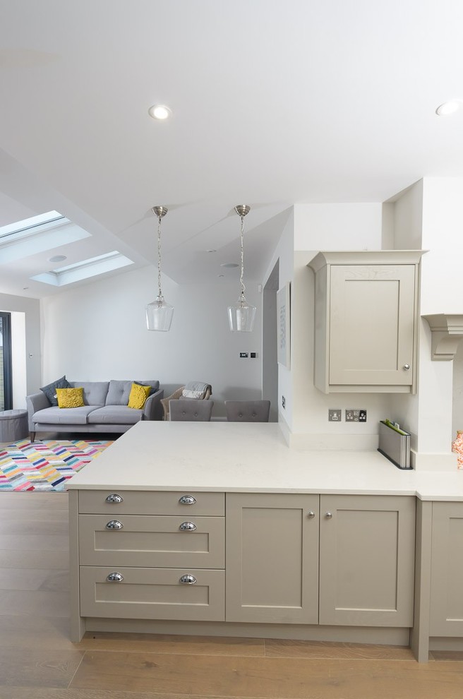 This is an example of a large contemporary galley kitchen/diner in Other with a belfast sink, shaker cabinets, beige cabinets, quartz worktops, white splashback, stone slab splashback, black appliances, light hardwood flooring, a breakfast bar, brown floors and white worktops.