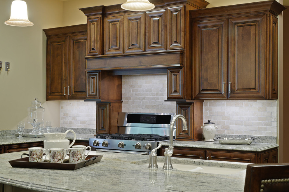 Cette image montre une cuisine traditionnelle en bois foncé avec un électroménager en acier inoxydable, un placard avec porte à panneau surélevé, une crédence beige et une crédence en travertin.