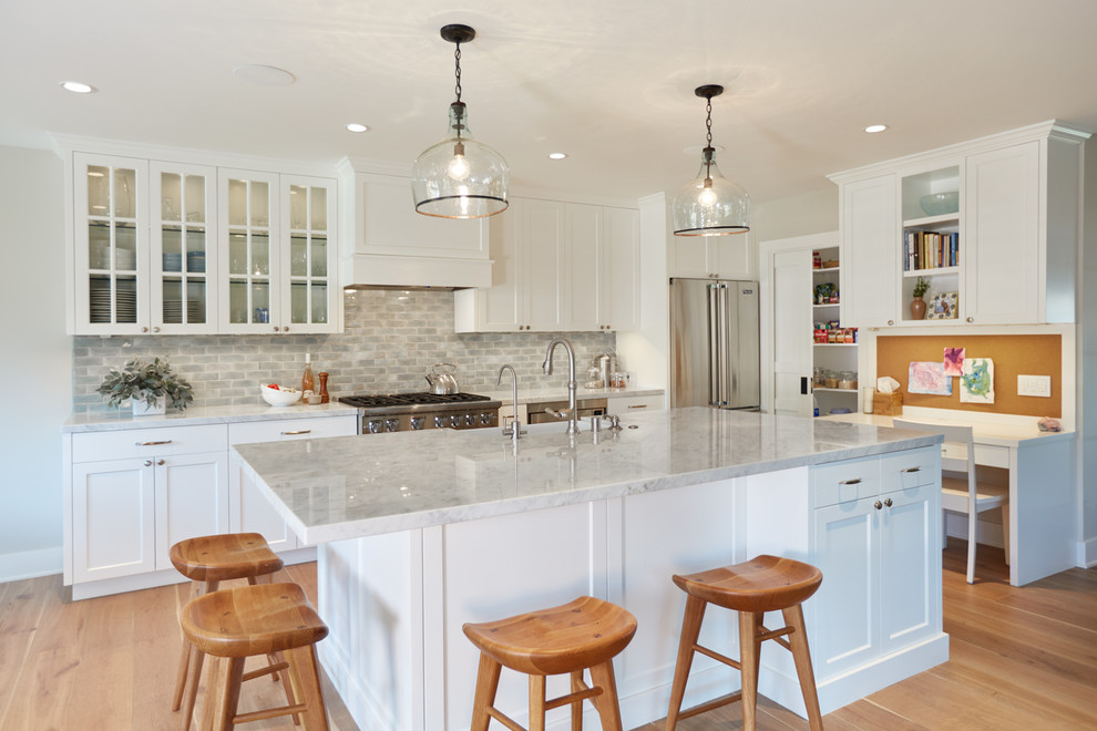 Large trendy l-shaped light wood floor eat-in kitchen photo in Los Angeles with a farmhouse sink, recessed-panel cabinets, white cabinets, marble countertops, gray backsplash, stone tile backsplash, stainless steel appliances and an island