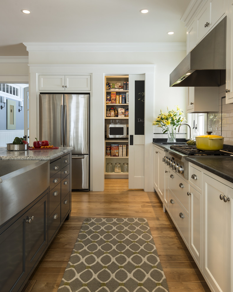 Example of a large classic l-shaped medium tone wood floor kitchen design in Portland Maine with a farmhouse sink, shaker cabinets, white cabinets, granite countertops, white backsplash, ceramic backsplash and stainless steel appliances
