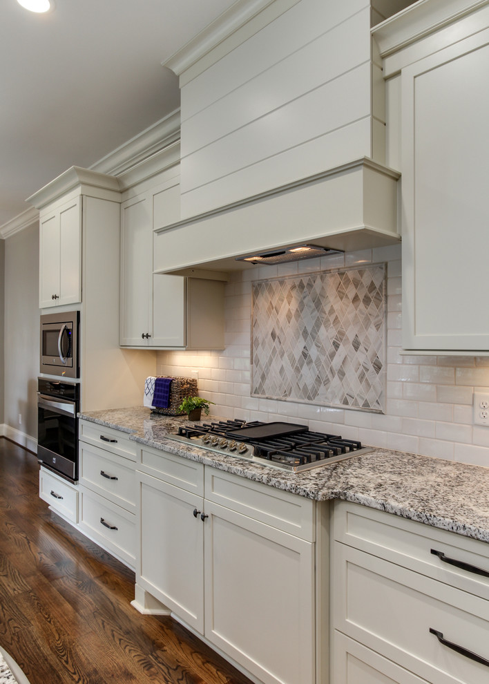 Photo of a medium sized classic l-shaped kitchen/diner in Raleigh with a single-bowl sink, recessed-panel cabinets, white cabinets, granite worktops, white splashback, ceramic splashback, stainless steel appliances, medium hardwood flooring and an island.